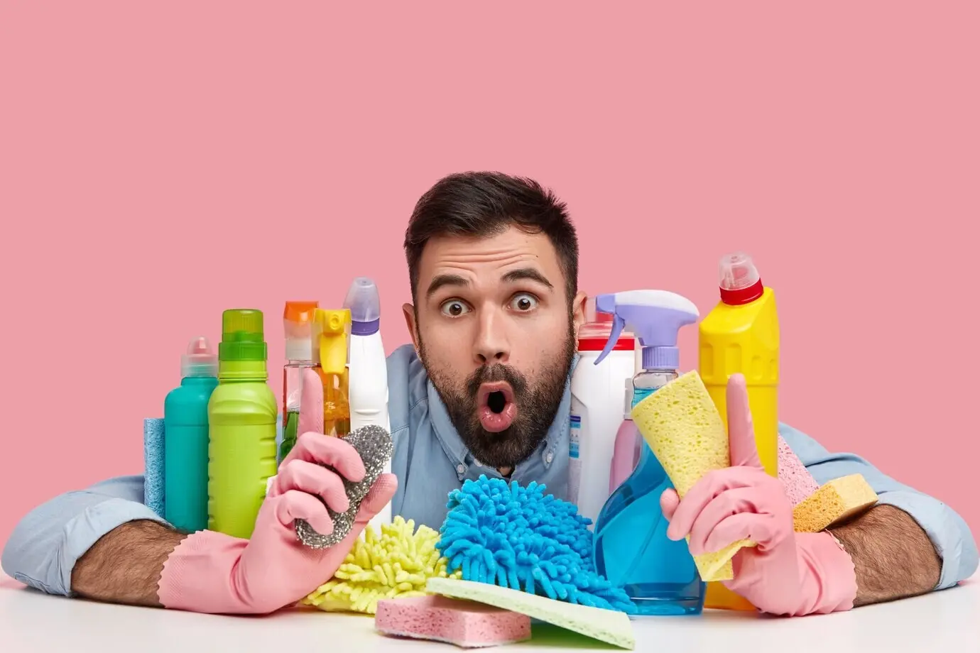 Hygiene- und Reinigungskonzept. Überraschter bärtiger Mann mit entsetztem Gesichtsausdruck sitzt am Tisch mit Reinigungsmittelflaschen und Schwämmen, mit heruntergeklappter Kinnlade.